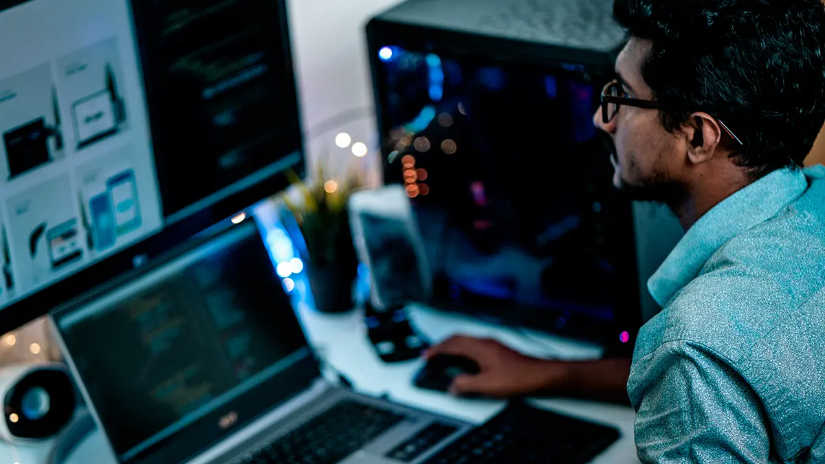 Pessoas trabalhando em computadores com múltiplas telas e códigos na tela, em um ambiente de escritório moderno, simbolizando a Plataforma End to End para Seguradoras