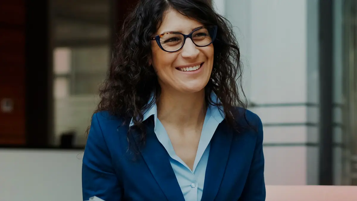Mulher adulta sorrindo em ambiente corporativo, vestindo blazer azul e óculos, em imagem com foco no atendimento e postura profissional. Seguro de Vida Universal no Brasil