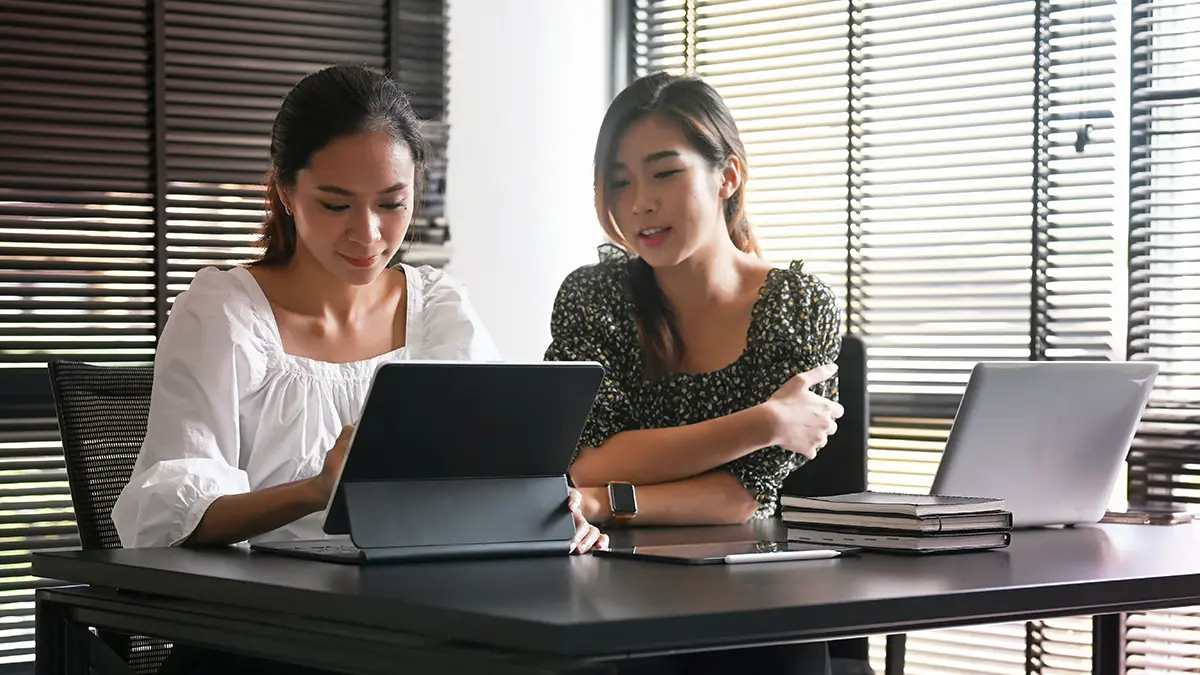 Duas mulheres em um escritório com janelas de venezianas, analisando informações em notebooks na mesa e discutindo um fluxo de trabalho com apoio de IA, EDI com IA para Corretoras