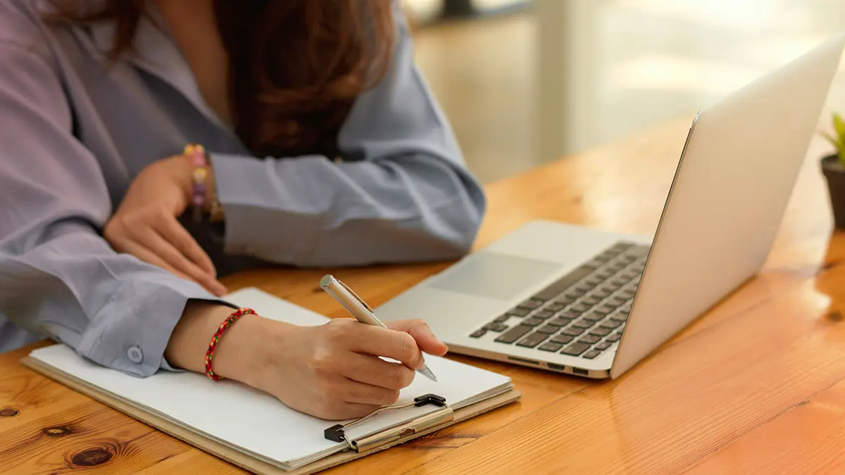Pessoa em escritório assinando documentos enquanto usa um notebook, com folhas e caneta sobre a mesa; cenário de Digitalização de processos com foco em organização e trabalho administrativo.
