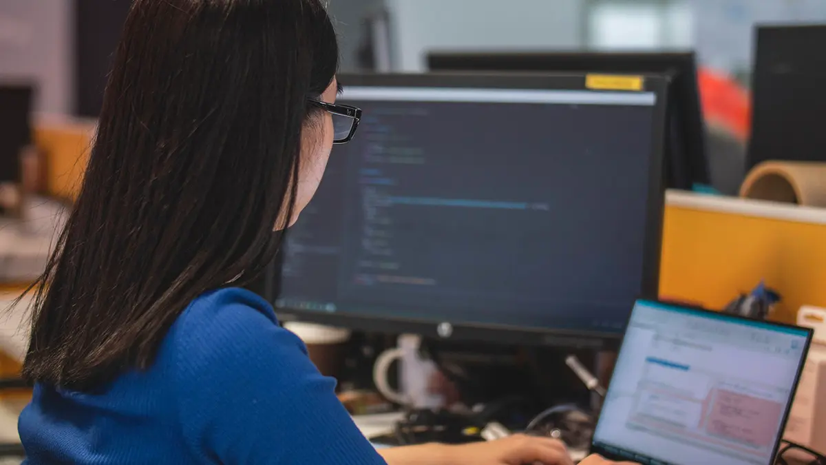Pessoa trabalhando em frente a dois monitores, analisando código e interfaces de software em um ambiente de escritório, com foco em programação e gestão de sistemas legados para Modernização de sistemas legados.
