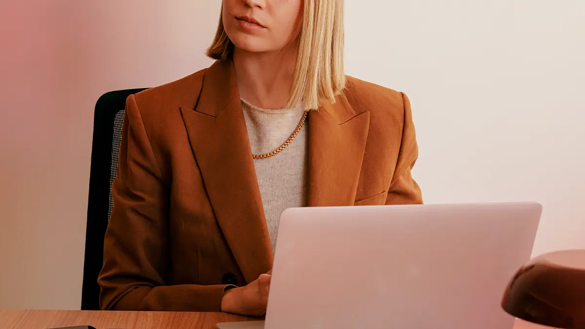 Integração de sistemas desafios: Profissional em ambiente corporativo, com blazer marrom e cabelo liso, utilizando notebook em reunião ao lado de outras pessoas, com foco em produtividade e colaboração.
