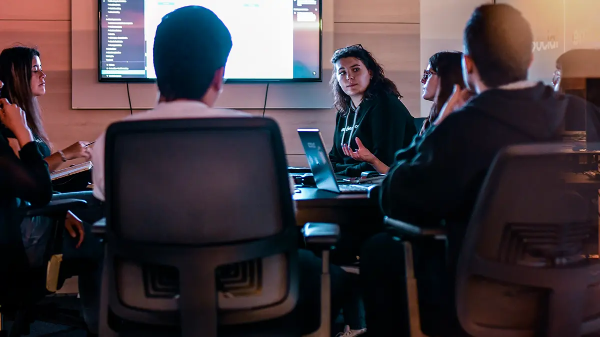 Equipe reunida em sala de reuniões com monitores exibindo gráficos e código, enquanto uma profissional apresenta ideias para Integração de sistemas empresariais durante discussão colaborativa