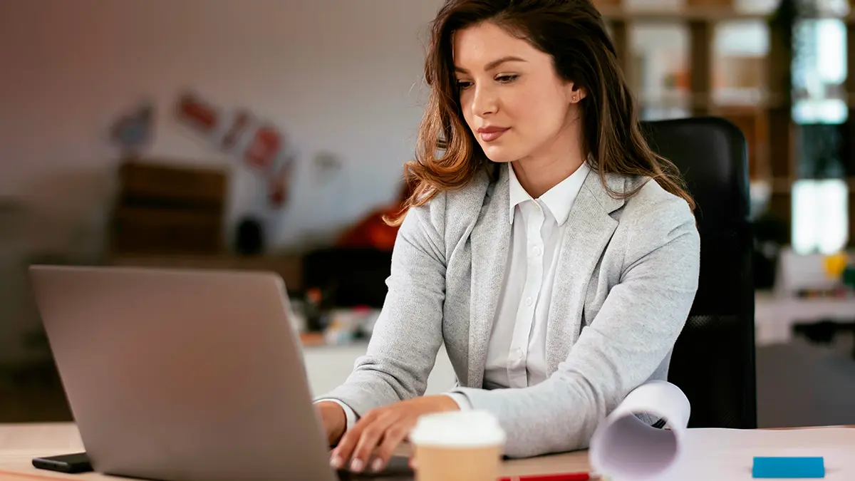 o que é API: Mulher trabalhando em um escritório, usando notebook e concentrada enquanto participa de uma reunião ou atividade profissional; ao fundo há objetos de trabalho.