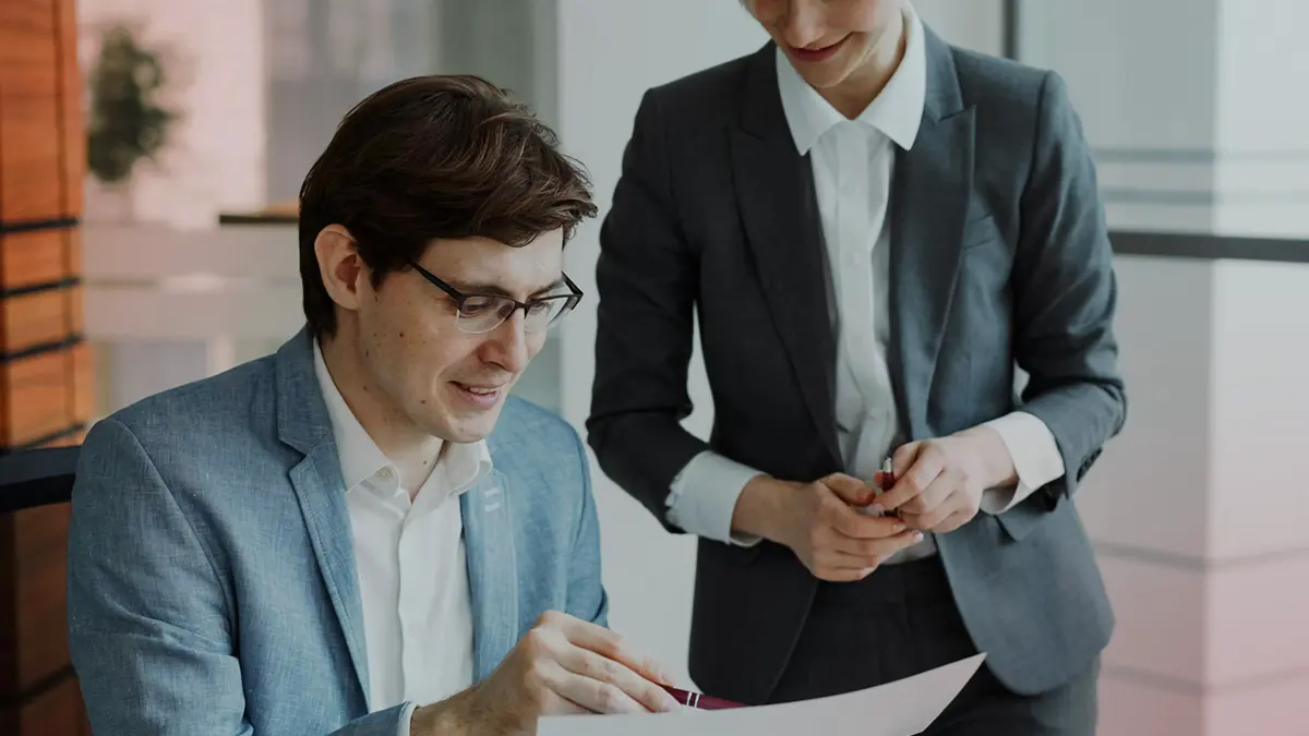 Dois profissionais em um escritório conversam e analisam documentos, enquanto um deles aponta com as mãos; a cena transmite orientação financeira e informações sobre o que é resseguro em contexto corporativo.