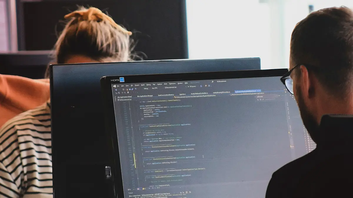 Profissionais trabalhando com programação em um computador, com código visível na tela, enquanto discutem soluções em ambiente corporativo, remetendo a outsourcing de TI para Seguradoras