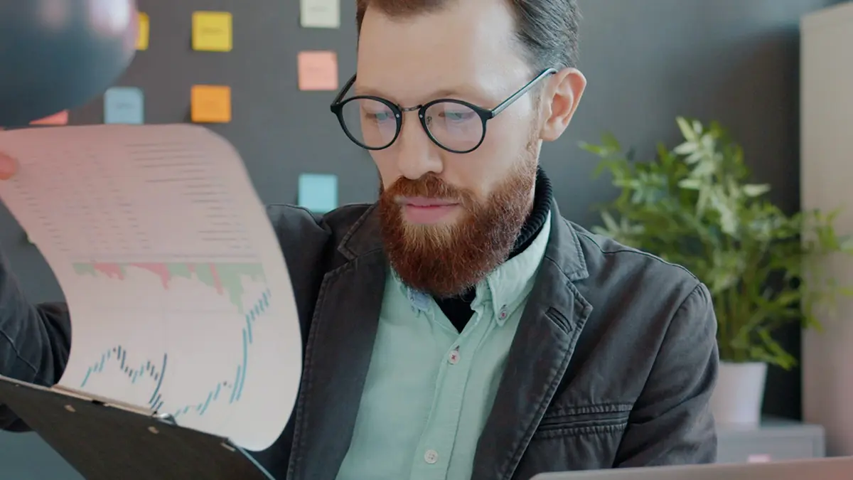 Analista com óculos e barba em ambiente corporativo, analisando gráficos e relatórios impressos ao lado de documentos com indicadores, representando qualidade de dados em seguradoras