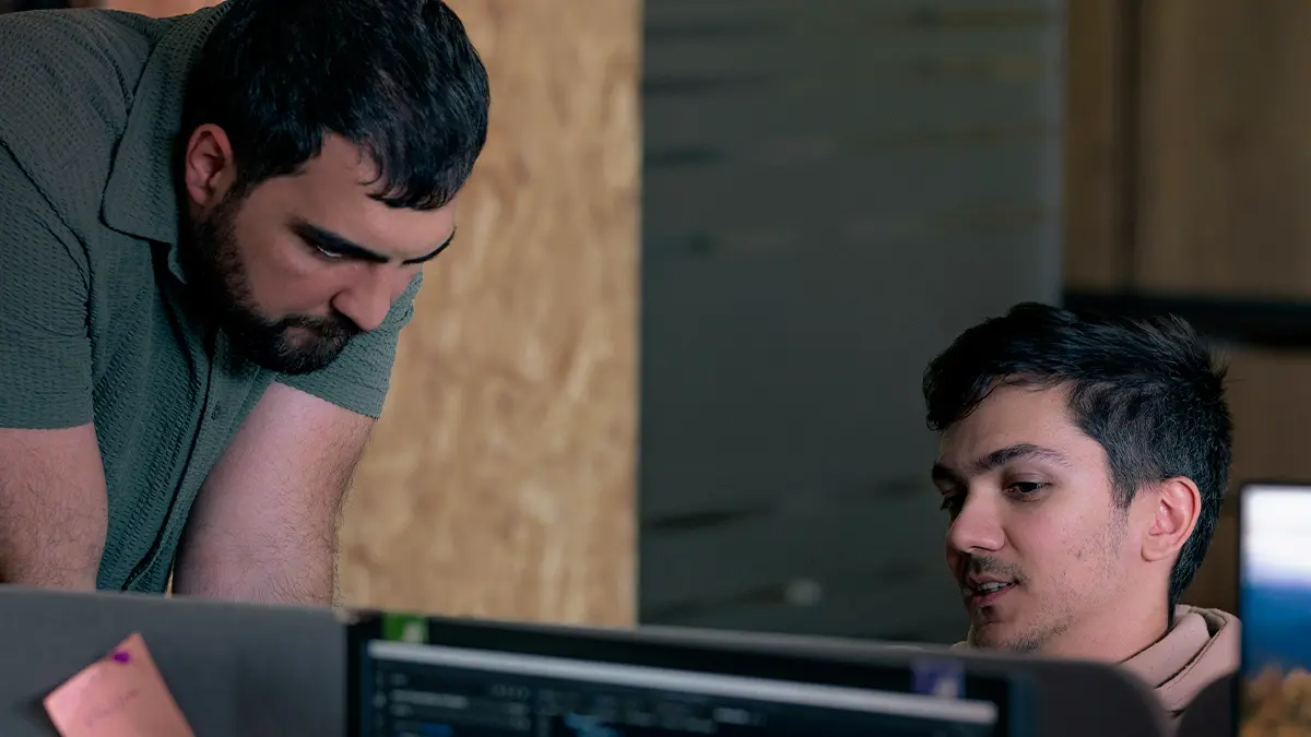 Dois profissionais analisam informações em um computador, em ambiente de escritório, enquanto um deles trabalha concentrado na tela e o outro observa a atividade, sugerindo segurança de APIs no setor de seguros.