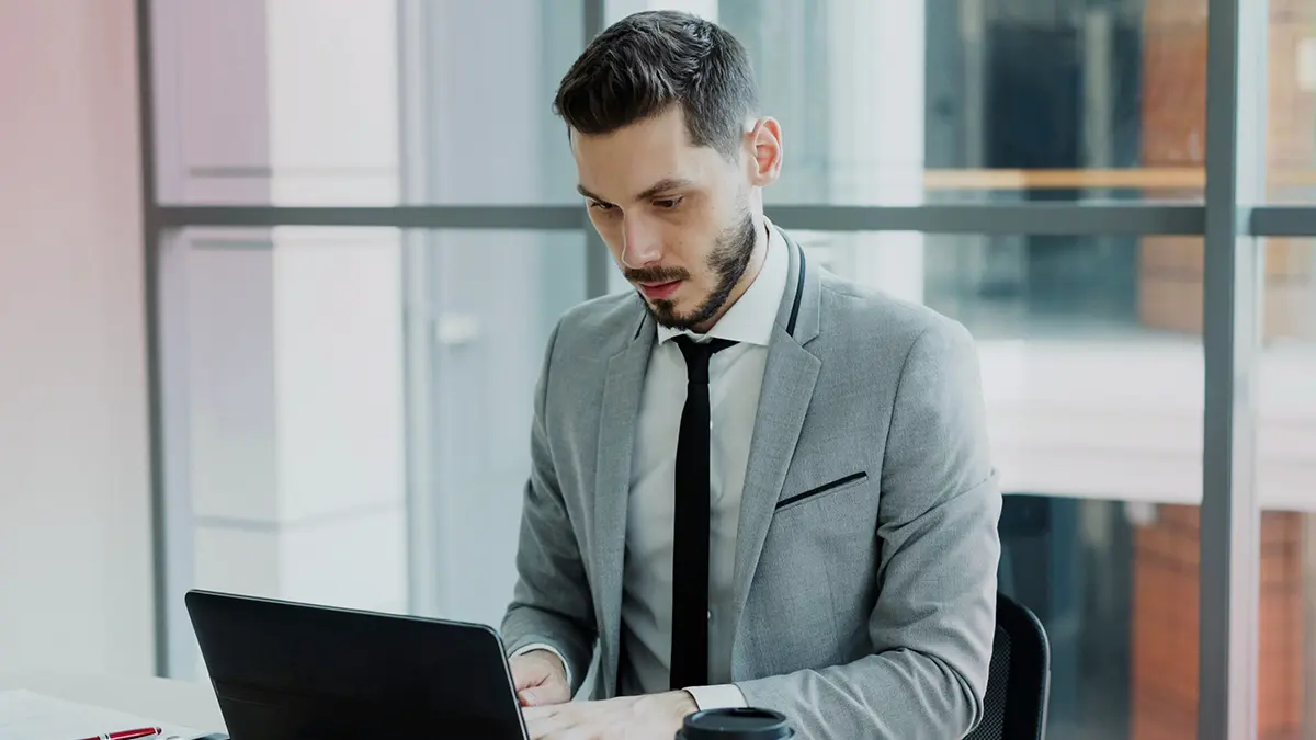 software para seguradoras: Homem de terno cinza em escritório, analisando informações no notebook e trabalhando em uma mesa com smartphone, em ambiente corporativo com janelas ao fundo.