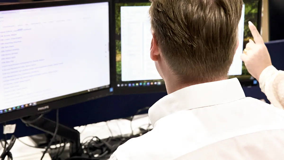Homem trabalhando em frente a monitores de computador e apontando para telas, em ambiente de escritório, com foco em processos e tecnologia para continuidade de negócios em seguradoras.