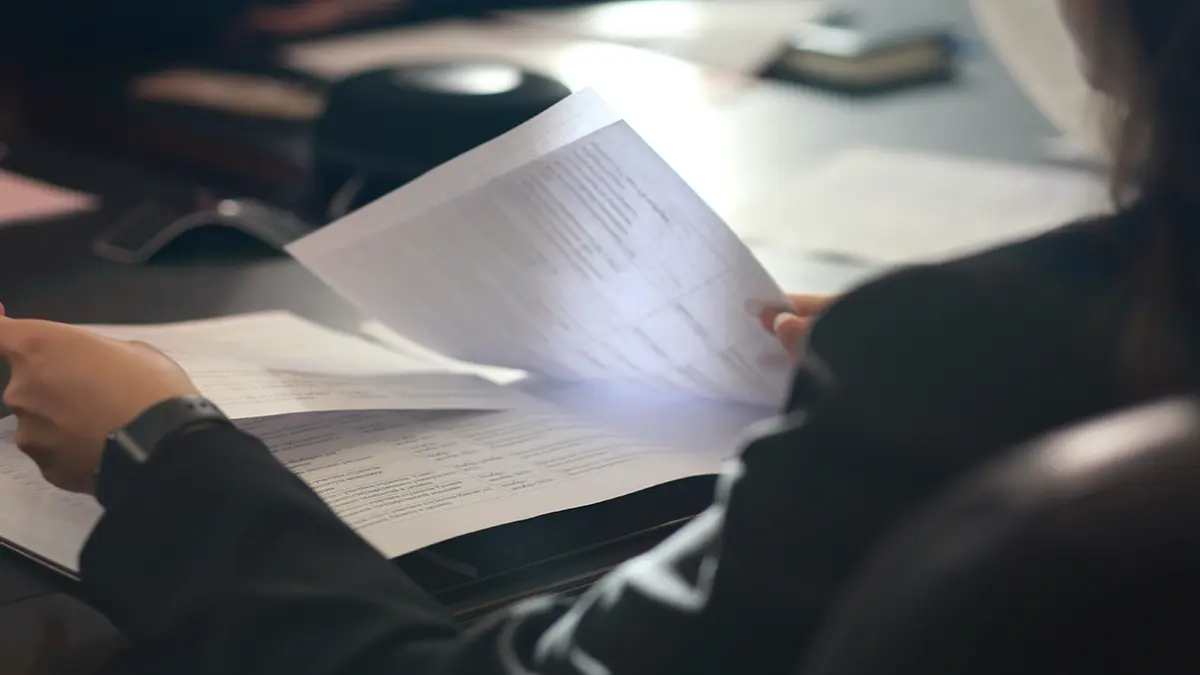 resolução CNSP 496: Pessoas analisando documentos e formulários em uma mesa, com papéis abertos e mãos virando páginas durante uma reunião de trabalho, em ambiente corporativo.