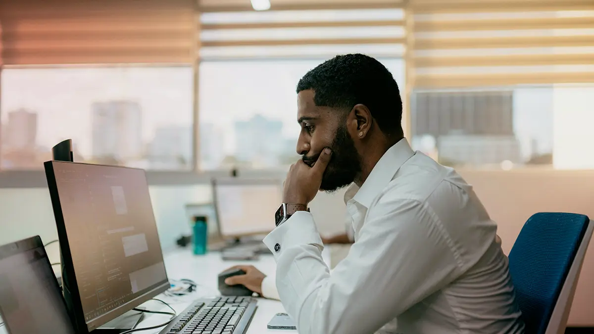 Homem trabalhando em um escritório, concentrado diante do computador com a mão no rosto, enquanto usa teclado e monitor. Resolução CNSP segurança cibernética em seguradoras496 sugerida no contexto de segurança digital.